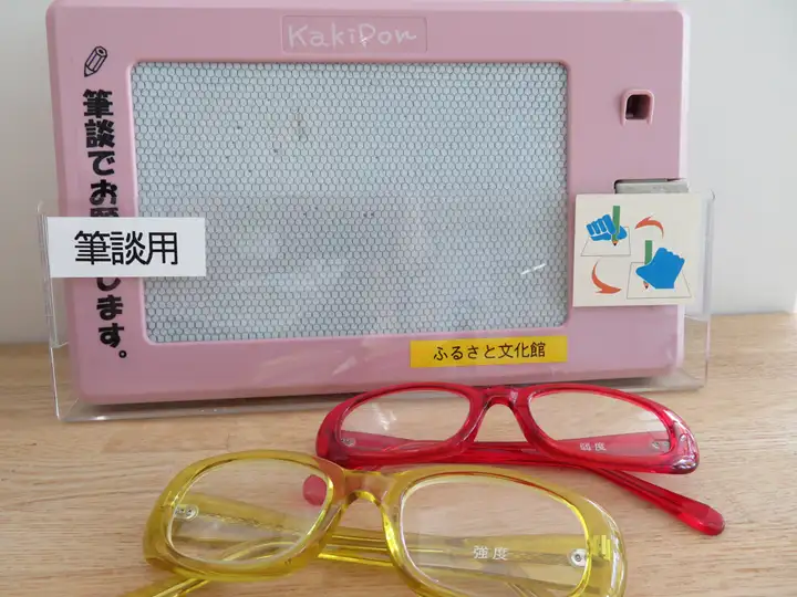 「筆談用」と掲示されたピンク色の磁気式筆談ボードと、赤色・黄色の老眼鏡
