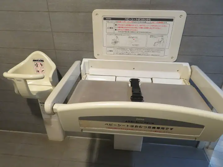 開いた状態の壁面設置型ベビーシート（おむつ交換台）と、その左横にある幼児用ベビーチェア